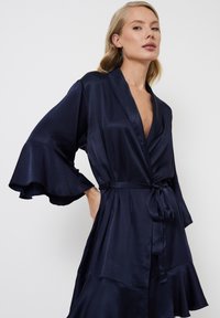 Robes satin marine avec des manches larges évasées, un décolleté en V, une taille cintrée et un détail d'ourlet superposé, affichant une texture lisse et brillante.