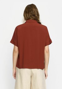 Rust-colored short-sleeve button-up shirt with a smooth texture and subtle sheen. Features a neat collar and rolled sleeves.