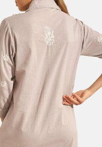 Tunique gris clair à manches longues, ornée de broderies florales blanches sur les épaules et le dos. Tissu doux et texturé avec un col.