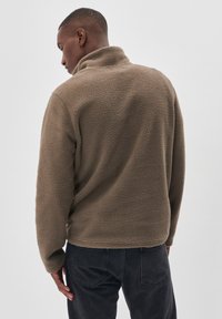 Fleece pullover i en dæmpet brun farve med høj krave, blød tekstur og minimalistisk design. Bæret sammen med mørke jeans.