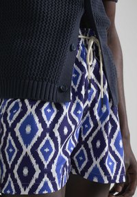 Cardigán de punto azul marino sobre pantalones cortos con estampado en azul y blanco que presentan un motivo de diamantes, cintura con cordón y detalles de botones negros.