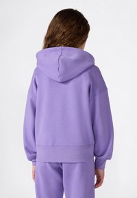 Persona con lunghi capelli castani indossa un felpa lavanda e pantaloni abbinati, di spalle contro uno sfondo bianco semplice.