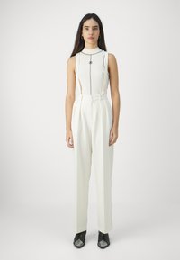 White sleeveless top with vertical accents and a logo, paired with high-waisted white trousers. Black heeled shoes complete the outfit.
