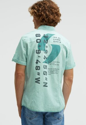 Mann mit lockigem Haar, der ein kurzärmeliges hellgrünes Hemd trägt, das bedruckte Zahlen, Text und eine große türkisfarbene Grafik auf der Rückseite zeigt.