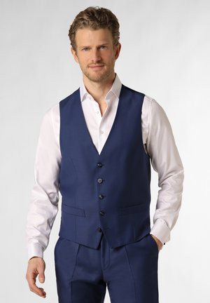 Mann trägt ein weißes Hemd, eine marineblaue Anzugweste und Hose, steht mit einer Hand in der Tasche vor einem neutralen Hintergrund.