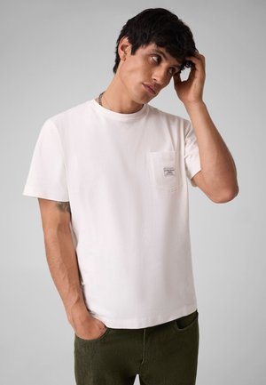 Ung mand iført en hvid lomme t-shirt og mørke bukser, rører ved sit hår med den ene hånd og har den anden hånd i lommen.