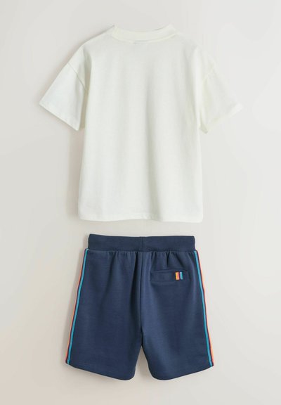 Chemise blanche à manches courtes associée à un short bleu marine avec des bandes latérales rouges, orange et bleues, et une petite poche arrière avec des bandes assorties.