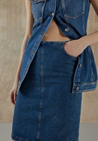 Džínová vesta se dvěma náprsními kapsami a viditelnými knoflíky, doplněná džínovou tužkovou sukní s boční kapsou a jemnou strukturou.