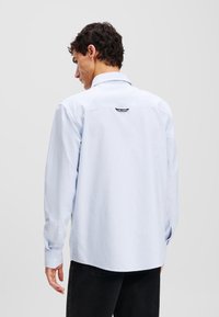 Light blue long-sleeve shirt with white stripes, button cuffs, and a rear logo tag. Cotton fabric with a smooth texture, casual fit.