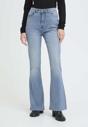 Femme portant un jean évasé taille haute bleu clair, un haut noir à manches longues et des bottines à talons noires, debout devant un fond uni.