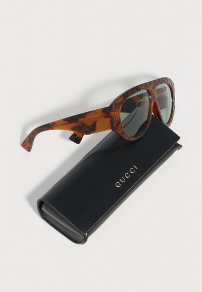 Lunettes de soleil Gucci à motif écaille de tortue avec verres foncés reposant sur un étui Gucci noir avec lettrage doré sur fond blanc.
