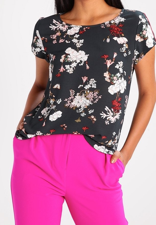 Femme portant un haut noir à manches courtes à motif floral, rentré dans un pantalon taille haute rose vif, les mains dans les poches.