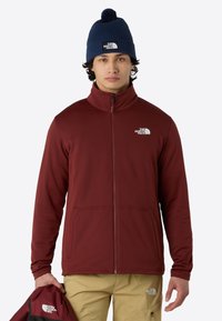 Giacca a zip bordeaux con colletto alto e due tasche frontali; presenta il logo The North Face. Berretto blu navy con pompon in cima.