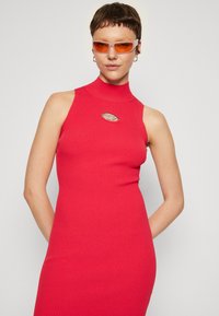 Abito aderente a coste di un vivace rosa con colletto alto e dettagli con foro. Modella che indossa occhiali da sole trasparenti, mostrando una trama liscia e un design senza cuciture.