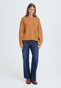 Pull en tricot couleur rouille avec un motif en losange texturé, associé à un jean bleu foncé et des bottines brunes. Le modèle se tient devant un fond clair.