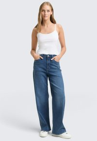 Damen-Jeans mit weitem Bein in Mittelblau und hoher Taille, kombiniert mit einem weißen Tanktop. Verfügt über ein klassisches Fünf-Taschen-Design und einen Knopfverschluss.