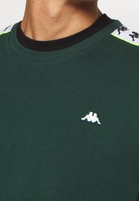 Grön sweatshirt med svart krage, med en vit Kappa-logotyp. Innehåller mönstrade axelaccent i vitt och svart med neongrön trim.