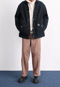 Svart knapp-up jacka över en krämfärgad hoodie, tillsammans med beige byxor med sidostråk och svarta klumpiga skor.