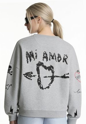 Donna con capelli biondi raccolti in una coda di cavallo indossa una felpa grigia con "Mi Amor,", una grafica a cuore e freccia e doodle a tema amore sulle maniche.