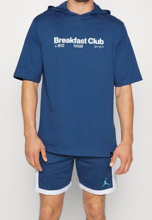 Mężczyzna ubrany w niebieską koszulkę z krótkim rękawem i kapturem z napisem „Breakfast Club” oraz pasujące niebieskie szorty sportowe z białym obszyciem i logo.