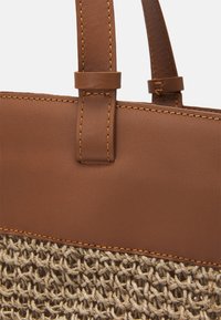 Sac fourre-tout en cuir marron avec fond en fibres naturelles tissées, doté de coutures marron et de poignées robustes. Design texturé et structuré.