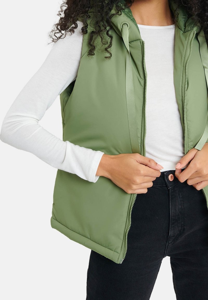 Femme portant un haut blanc à manches longues, un gilet matelassé vert olive et un jean noir taille haute, fermant la fermeture éclair de son gilet.