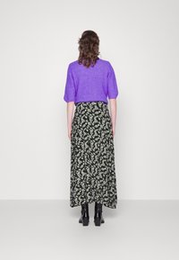 Femme aux cheveux bruns bouclés, portant un pull violet, une jupe maxi noire à fleurs et des bottines à talons noires, se tenant face à un mur blanc.