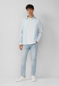 Lichtblauwe lange mouwen shirt met kleine patronen, vooraan geknoopt, gecombineerd met lichte jeans en witte sneakers. Model staat recht vooruit.