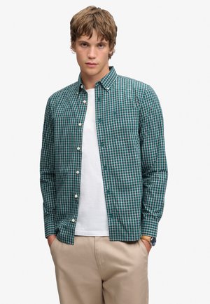 Ung mand iført en grøn- og hvidternet skjorte over en hvid T-shirt og beige bukser, stående med hænderne i lommerne.