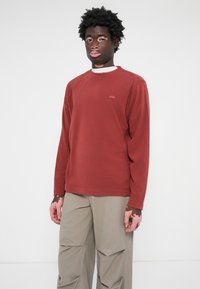 Röd sweatshirt med långa ärmar och rundad krage. Tillverkad av mjukt tyg, med en liten logotyp på bröstet, parat med beiga cargobyxor.