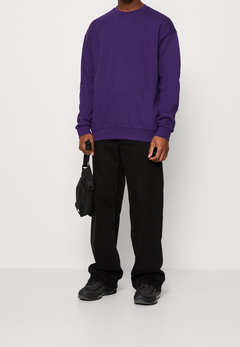 Felpa viola con collo a giro, polsini e orlo a coste, abbinata a pantaloni neri a gamba larga e sneakers neri. È in mano una piccola borsa nera.