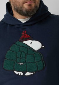 Granatowa bluza z kapturem z pluszową, haftowaną postacią w zielonym pikowanym płaszczu i czerwonym kapeluszu. Projekt zawiera wyraźne kontury i faktury.