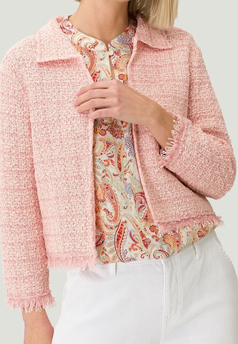 Giacca in tweed rosa con colletto strutturato, bordi sfrangiati e maniche a tre quarti, indossata sopra una blusa a motivi e una cintura bianca.