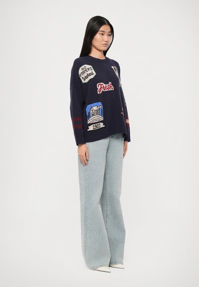 Navy sweater with embroidered patches, including graphics and text. Features red and white stripes on sleeves. Paired with light blue wide-leg jeans.