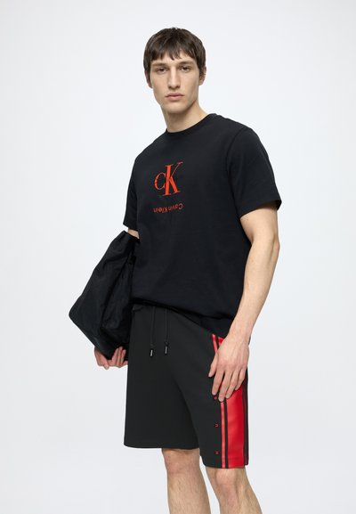 Мъж, облечен в черна тениска Calvin Klein и черни шорти с червени ивици, държащ черно яке на бял фон.