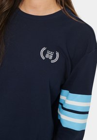 Felpa navy con collo a girocollo a costine, caratterizzata da un logo ricamato bianco e da strisce alternate blu e bianche sulle maniche.