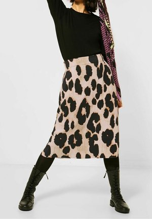 Kvinde iført sort langærmet top, beige midi-nederdel med leopardprint, sorte strømpebukser og sorte snørebånds-kampsko, stående med benene spredte.