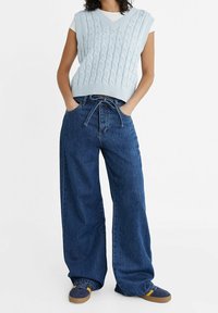 Hellblauer, kabelgestrickter, ärmelloser Pullover kombiniert mit locker sitzenden dunkelblauen Jeans mit Kordelzugbund und Sneakers.