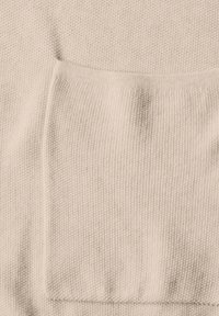 Ljust beige stickad tyg med en texturerad väv. Har en stor, fyrkantig framficka. Mjuk och mysig utseende.