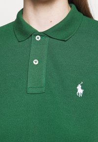 Grön polo skjorta gjord av strukturerad bomull, med en klassisk krage, tre knappar och en vit broderad logotyp på vänster bröst.