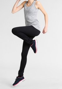 Graues sportliches Tanktop, schwarze enganliegende Hose und marineblaue Laufschuhe mit pinken Akzenten. Glattes Material, stromlinienförmiges Design, Bein angehoben.