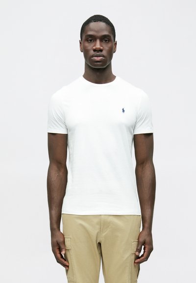 Fehér pamut rövid ujjú póló környakú kivitelben, a mellkason egy kis tengerészkék logóval. Bézs cargo nadrággal párosítva.