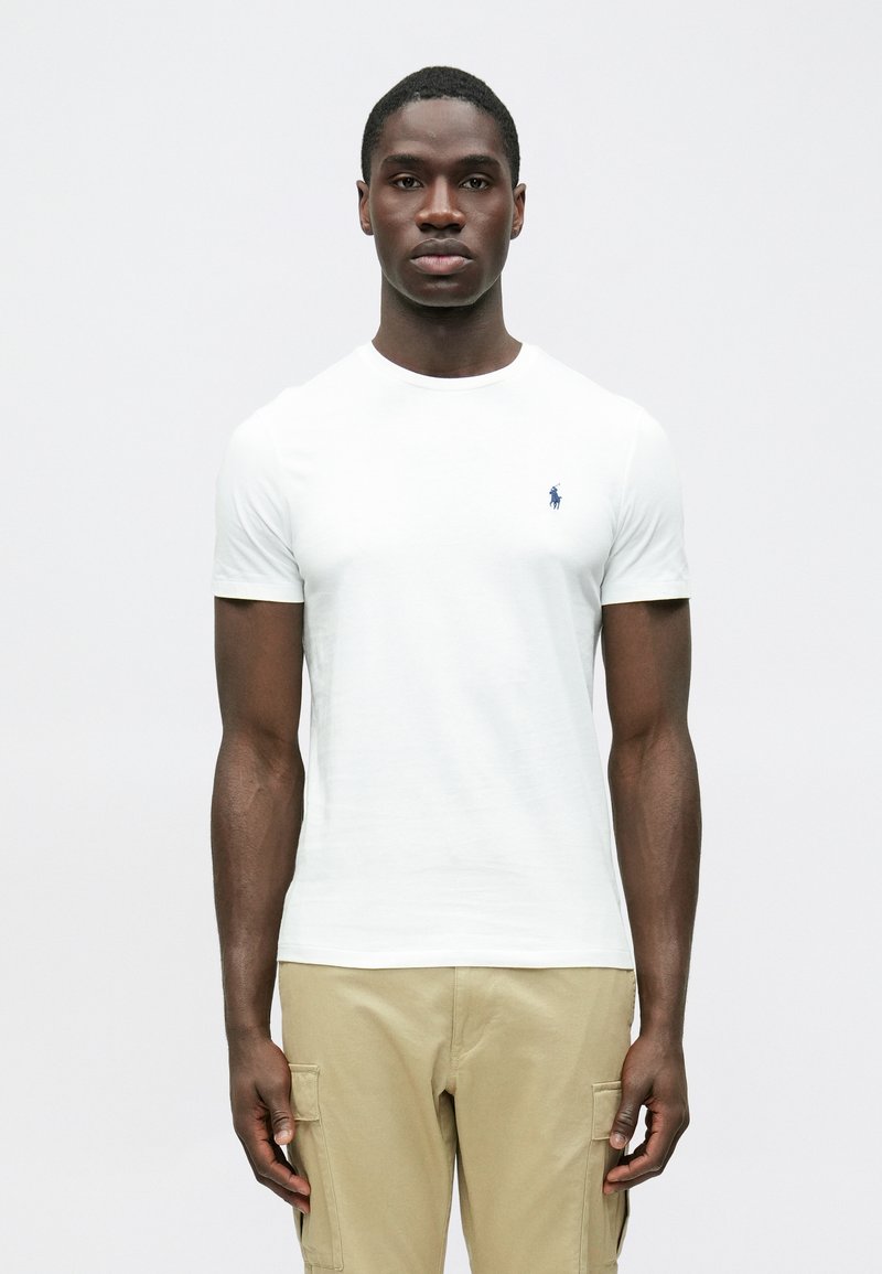 T-shirt bianca in cotone con maniche corte e scollo tondo, con un piccolo logo blu navy sul petto. Abbinata a pantaloni cargo beige.