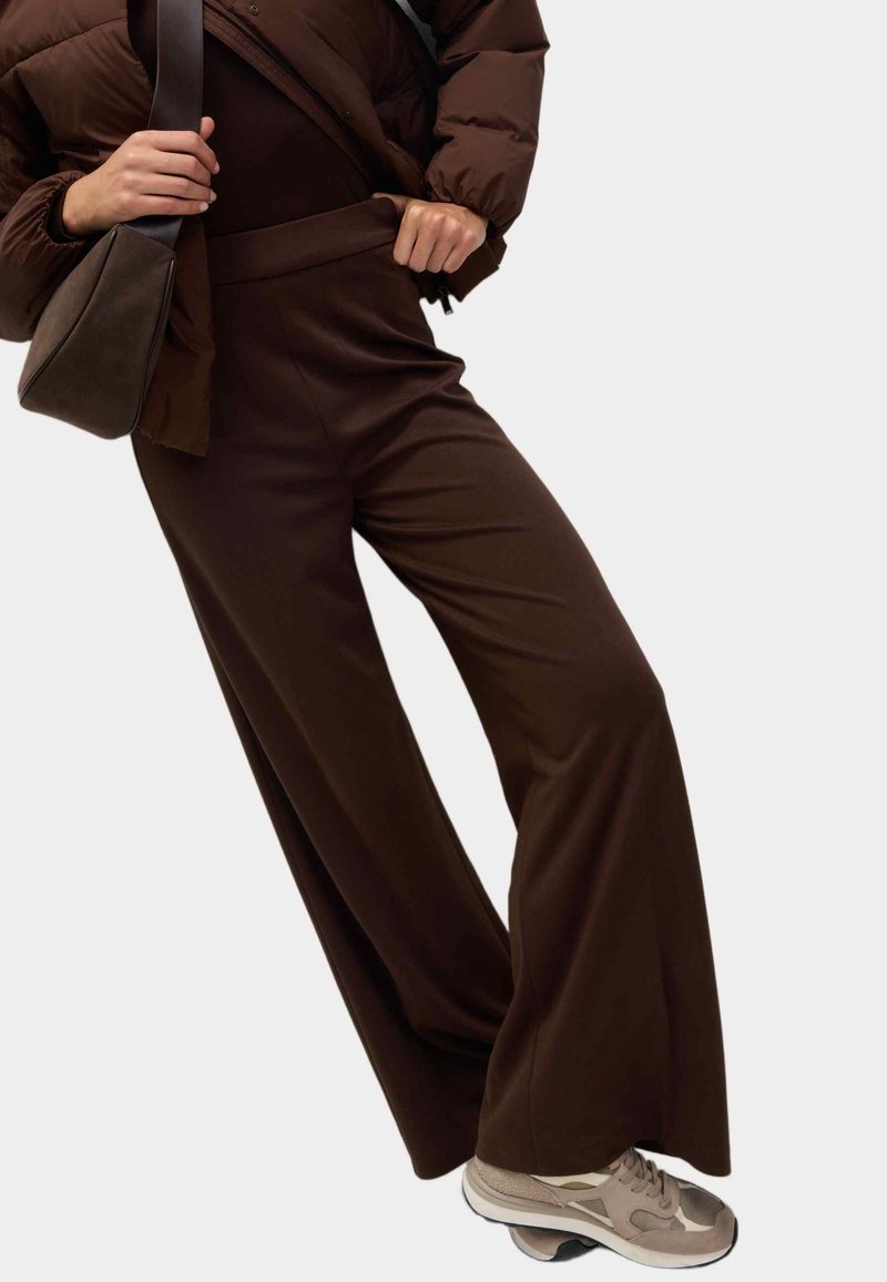 Personne portant un pantalon large marron foncé, des baskets marron clair, une doudoune marron et tenant un sac bandoulière marron foncé.