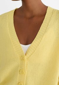 Yellow knitted cardigan with a deep V-neck, ribbed collar, and four yellow buttons. The texture appears soft and lightweight.