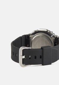 Montre avec bracelet en caoutchouc noir, boucle en métal argenté, surface texturée et fond métallique rond. Résistante à l'eau.