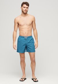 Blauwe patroon zwembroek met een elastische tailleband en trekkoord, met een comfortabele pasvorm en een korte broeklengte. Geddragen met zwarte teenslippers.