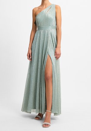 Kobieta ubrana w błyszczącą, jasnooka zieloną suknię na jedno ramię z wysokim rozcięciem na udzie i szpilki z paskiem wokół kostki, stojąca na tle jednolitego tła.