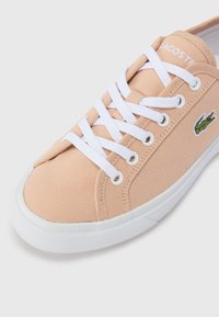 Lys ferskenfarget kanvas sneaker med hvite snørebånd og gummisåle. Har en grønn brodert krokodillelogo på siden. Flat, rund tå.