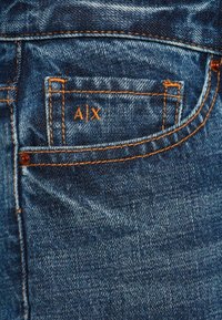 Denim-farkut, joissa on oranssit tikit ja A|X-logo brodeerattuna pienessä sisätaskussa.